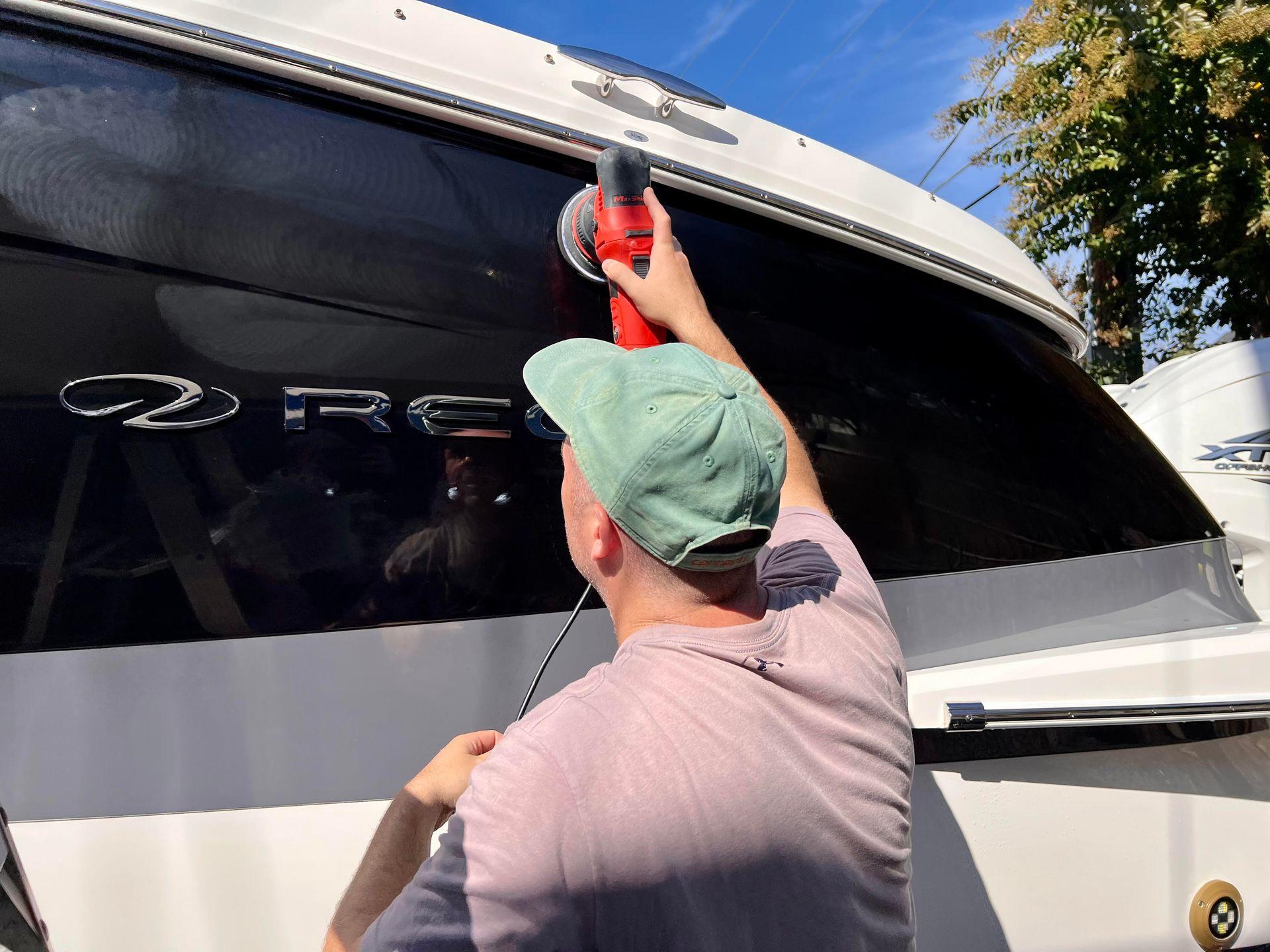 Person polishing the side of a boat with an electric buffer under a blue sky.